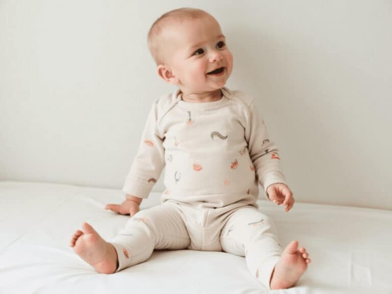 Baby sitting on bed in cream pajamas with animal print, smiling outdoor, childhood innocence, baby comfort, adorable infant image.