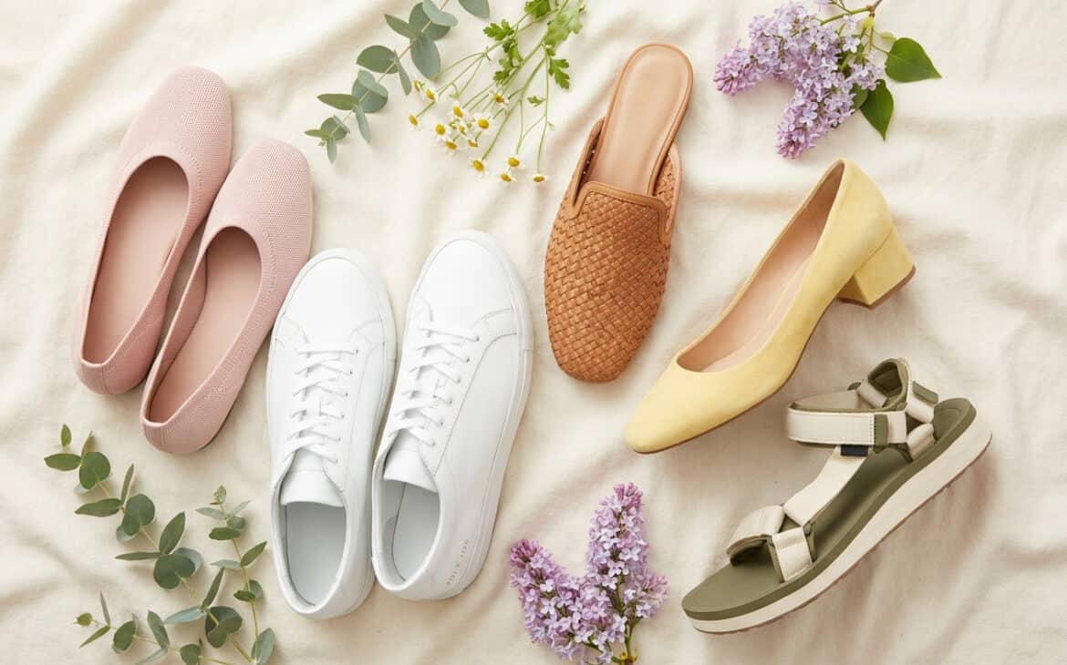 A flat-lay of stylish spring shoes: knit flats, white sneakers, a tan mule, a block-heel pump and a sport sandal