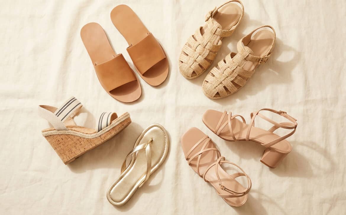 Flat-lay of five summer sandal styles: leather slide, fisherman sandal, cork espadrille wedge, metallic thong, and strappy block heel